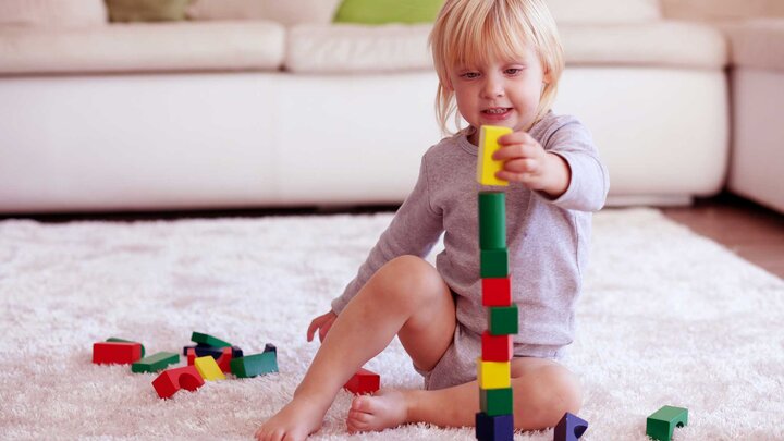 A young child with blonde hair, sitting on a light colored carpet, stacking colorful blocks. 