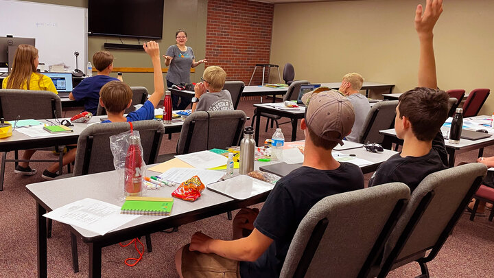 A tan and brick walled classroom setting. Instructor speaking to a group of students. Lots of school supplies and snacks sitting on tables. Photo taken from the back of the room. 