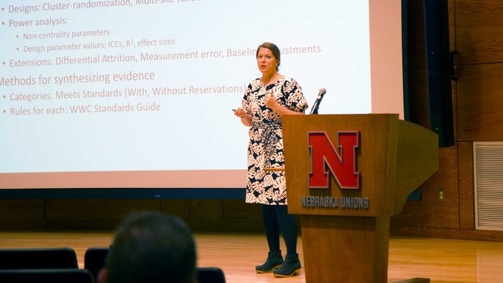Individual standing near a wooden podium in front of a large presentation screen. Individual is wearing a black and white floral dress. 