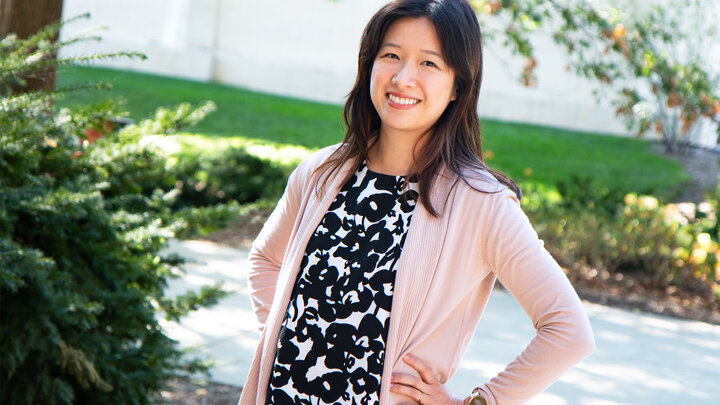 Individual with long dark hair standing outside, smiling and posing for a photo. Individual pictured is wearing a black and white floral shirt, covered by a light pink sweater. 