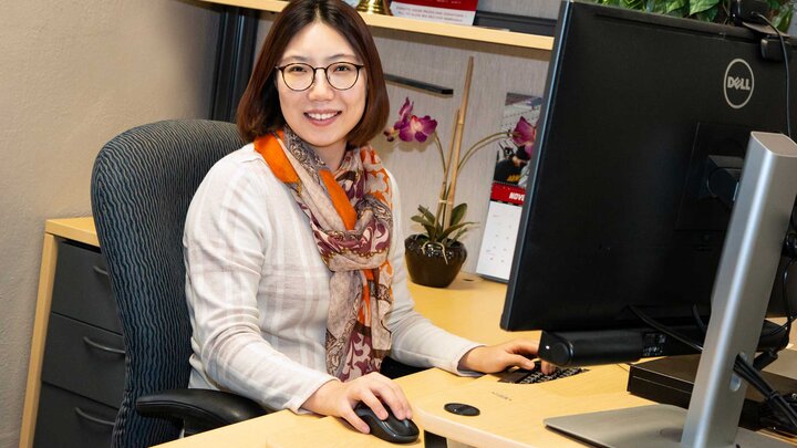 Professional office setting. An individual wearing a tan sweater and orange scarf smiles for a photo while sitting at a computer. 