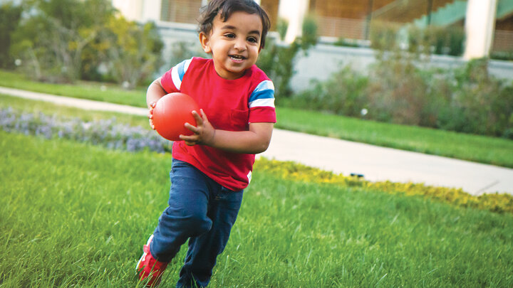 Small child playing on the grass outside. Child is wearing a red shirt with jeans, and is running with a small orange ball. 