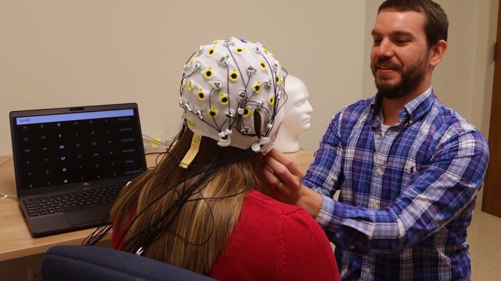 Two individuals in an office setting. One person, wearing a blue shirt is placing a medical device on a head. The device has many cords attached. 