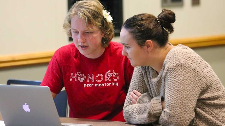 Two individuals sitting at a table looking at a computer. One is wearing a read shirt that reads 