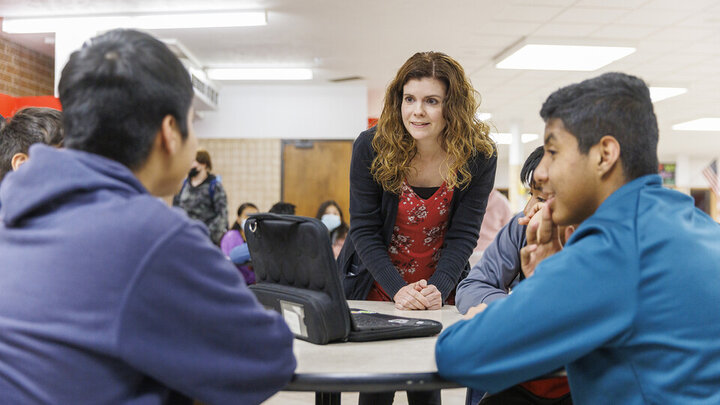 School setting, one individual standing, talks to two individuals, sitting at a table. 