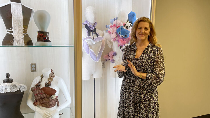 Individual in a black dress, stands holding a glass star-shaped award in front of a display case. The display case features colorful and unique garmets. 