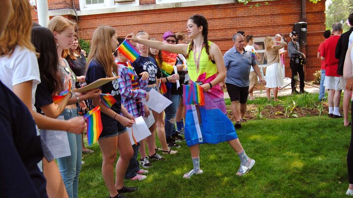 A large group of youth stand outdoors. One is passing out colorful flags to the group. 