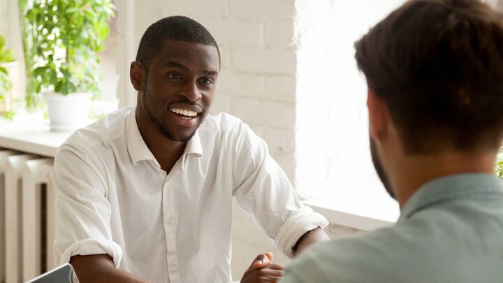 One individual, smiling, sitting outside on a porch or balcony having a conversation with another individual who's back is to the camera. 