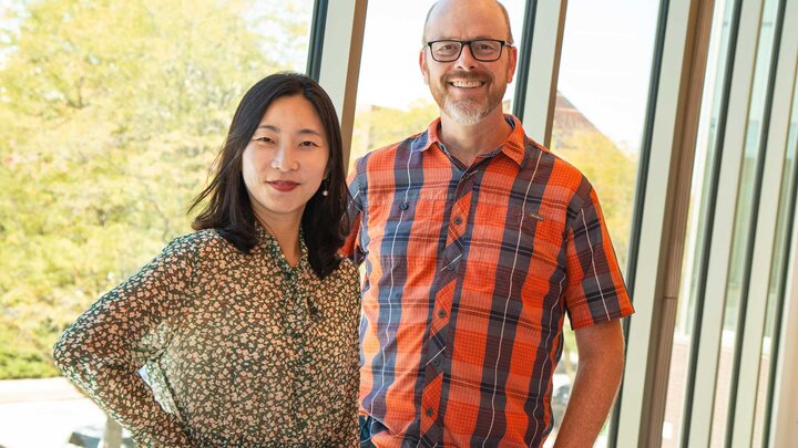 Two adults, one wearing a green floral shirt and the other wearing a red and blue plaid shirt, stand smiling in front of large windows. 