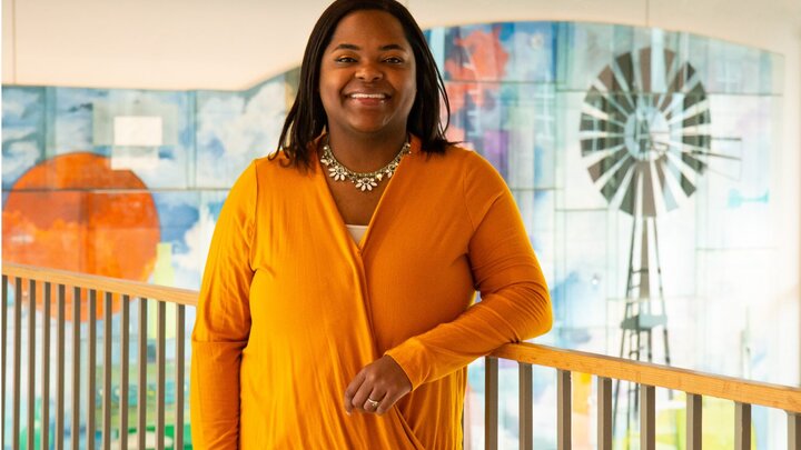 Jasmin Smith, wearing an orange shirt, stands in front of a colorful mural inside of Carolyn Pope Edwards Hall and smiles for a photo.