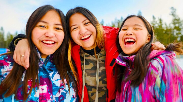 Three Indigenous youth wear bright clothing and smile for a photo while standing outdoors. 