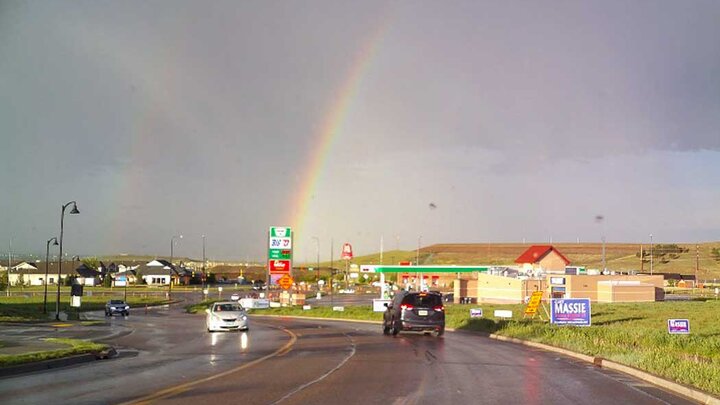 A rainbow shines over a highway with a gas station and other community buildings on each side. 