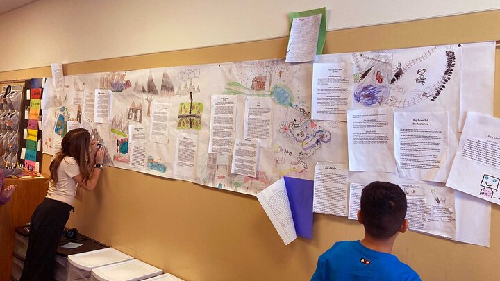 Two students stand at a wall filled with papers of sketch drawings and text. 