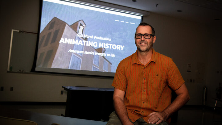 Michael Burton sits in a dark classroom in front of a large screen displaying the Animating History webpage. 