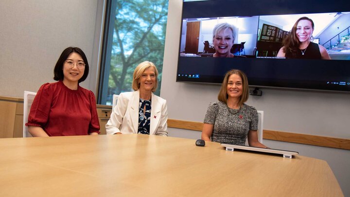Three people dressed in business clothes sit at a wooden desk. Behind them is a large monitor featuring two other individuals connecting virtually.