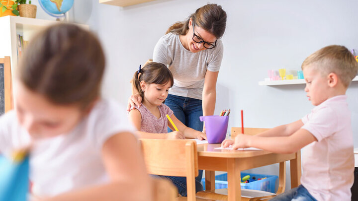 teacher helping a young girl in a classroom as other students also work at their desks