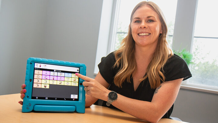 Ciara Ousley sits at a table smiling while pointing to an AAC tablet