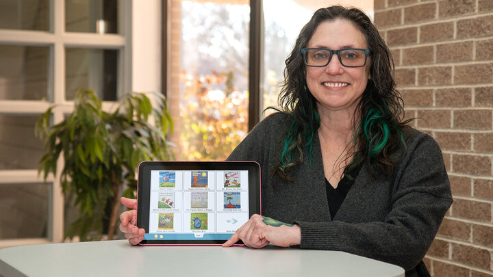 Birdie Ganz stands by a table while pointing at an AAC device and smiling for the photo