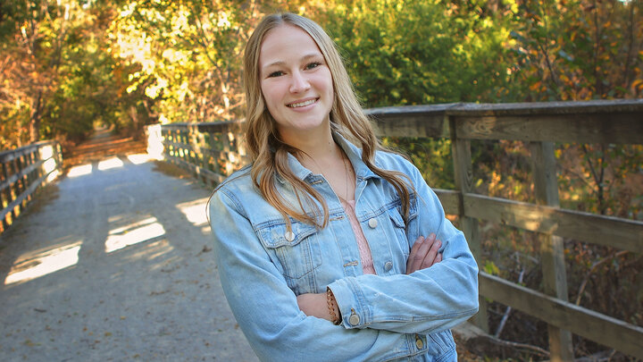 Sela Rikli smiles with arms crossed standing on a trail bridge with sun shining through the trees in the background