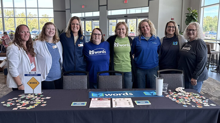 eight members of the WORDS team pose for a photo behind a table with information about the WORDS Project at the NeMTSS conference