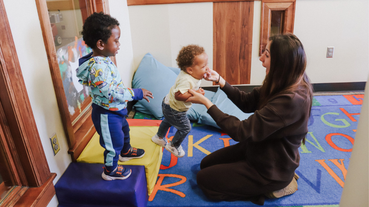 an educator kneels on the floor while playing with two young children on a mat at the University of Nebraska-Lincoln Children's Center