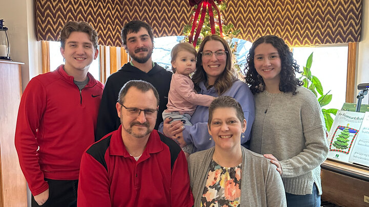Jennifer Boldt (front right) poses for a family photo with five other adults and a young child in front of a Christmas tree and large window in the background