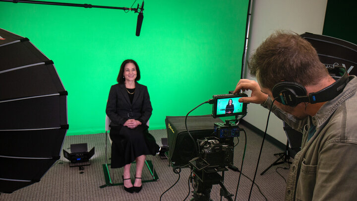 Film studio setup with a green screen, camera, lighting equipment, and a seated person being recorded.
