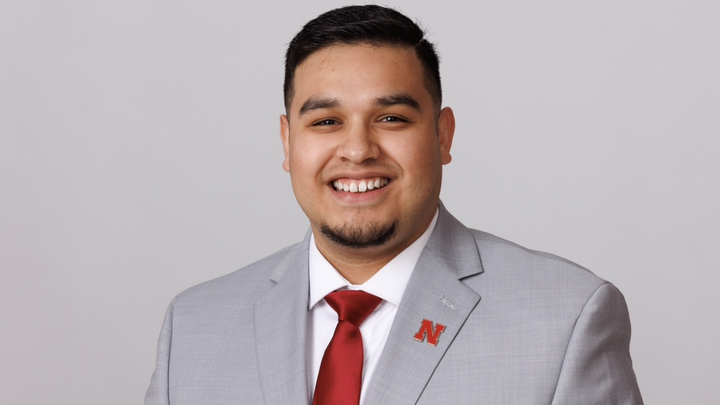 Jhovany Millan headshot wearing light gray suit coat with white shirt and red tie, red N pin on left lapel; light gray photo background