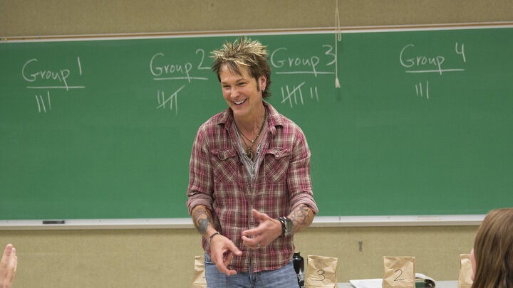 John Maag smiles while standing at the front of a classroom with a large green chalkboard and a table with brown paper bags in the background