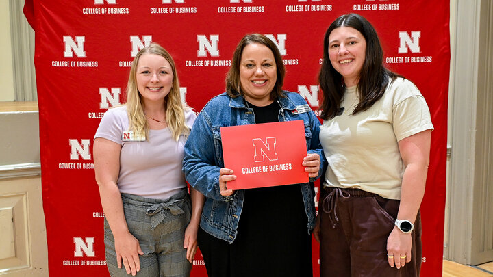 Courtney Eitzmann, AnnMarie Gottner and Abbie Reynolds pose for a photo in front of a red photo backdrop with white N College of Business lockups pattern while AnnMarie holds a red folder with an N College of Business on it