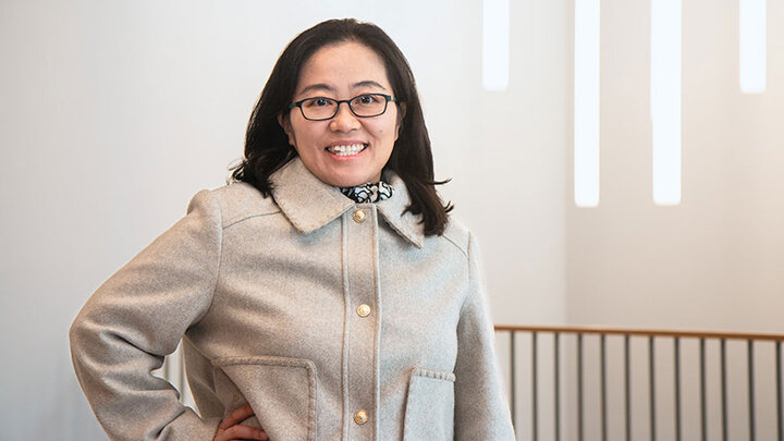 Sungeun Kang stands with her right hand on her hip wearing a cream-colored jacket buttoned up; light-colored walls, gray and wood railing and four vertical lights in background