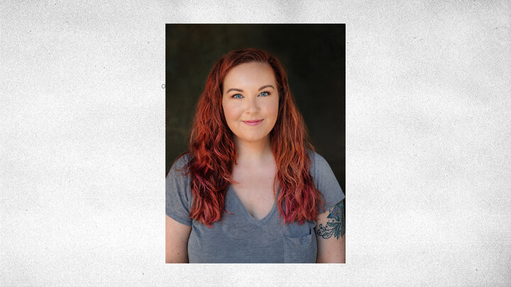 Smiling woman with red wavy hair wearing a grey shirt, set against a dark background, centered on gray textured background