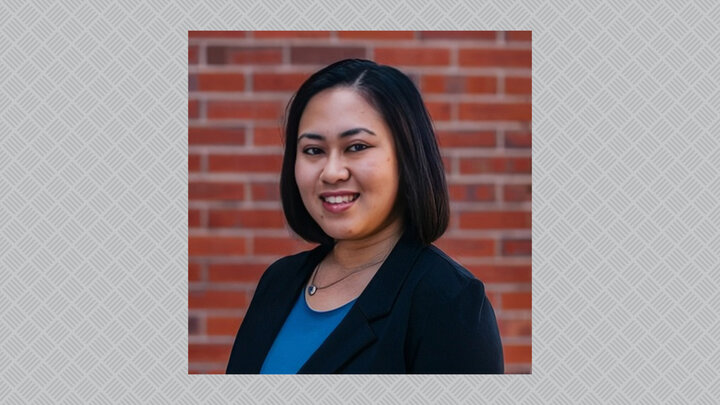 Smiling woman in blue shirt and black blazer, standing against a brick wall background; centered over gray background with crosshatch design