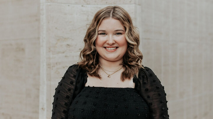 Smiling woman with wavy hair wearing a black dress against a neutral stone background.