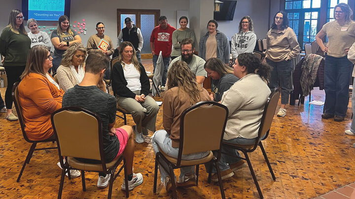 Group of people seated in a circle discussing, surrounded by others standing in a large room.