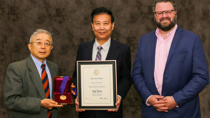 Three individuals dressed in business attire pose for a photo. One individual, in the center holds a large plague, and another holds a wooden box displaying a gold medal. 