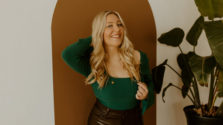 Allie Gotchall stands in front of a wall painted with geometric shapes, and next to a tall green plant. 