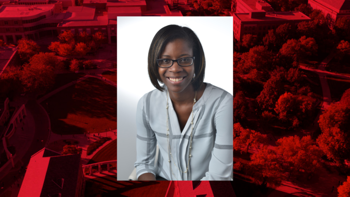 Professional headshot on red background. Individual pictured is wearing a light blue shirt, long necklace and eye glasses. 