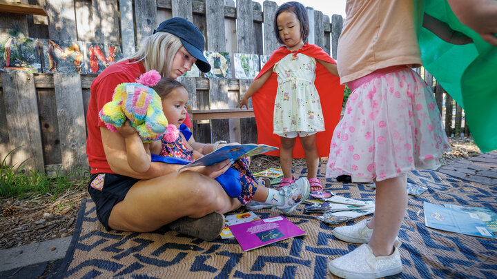 One adult, sits outside reading a book to three children. 