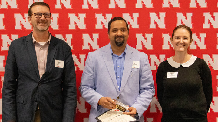 Three individuals in professional attire standing and posing for the photo in front of a red background with white N's.