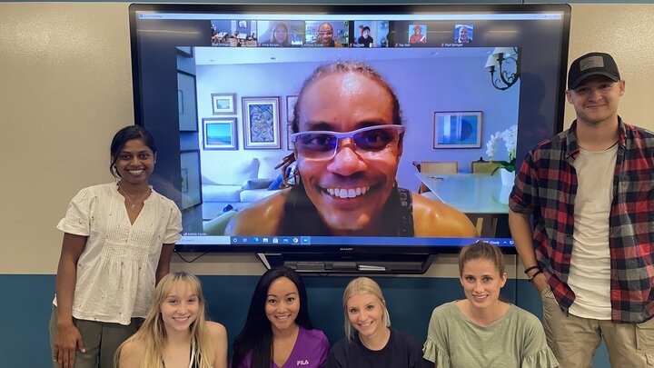 Six individuals standing in front of a blue and gray wall with a tv hanging on it. An additional person is on the tv screen smiling.