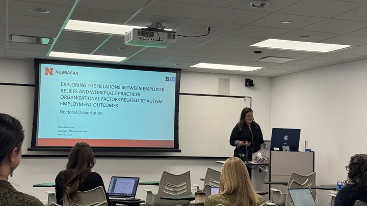 Woman presenting a doctoral dissertation in a classroom with attendees seated and attentive.