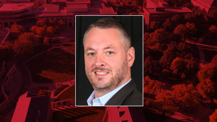Man with short hair and beard in formal attire, framed over a red-tinted aerial view of a campus.