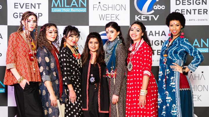 Seven individuals pose on a red carpet, wearing hand crafted looks that were showcased at Omaha Fashion Week. 