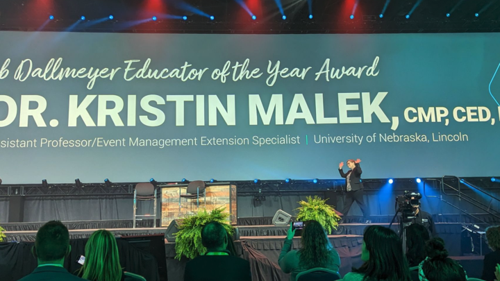 Kristin Malek walks across a large stage with a screen displaying the educator of the year award information. 