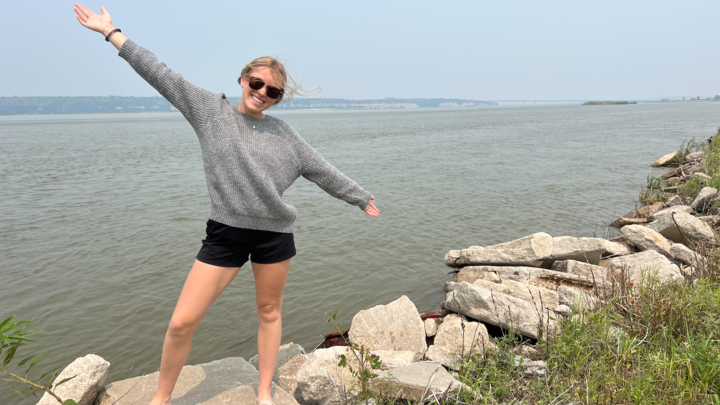 Maycee Quick smiles and spreads her arms out while standing in front of a large river. 