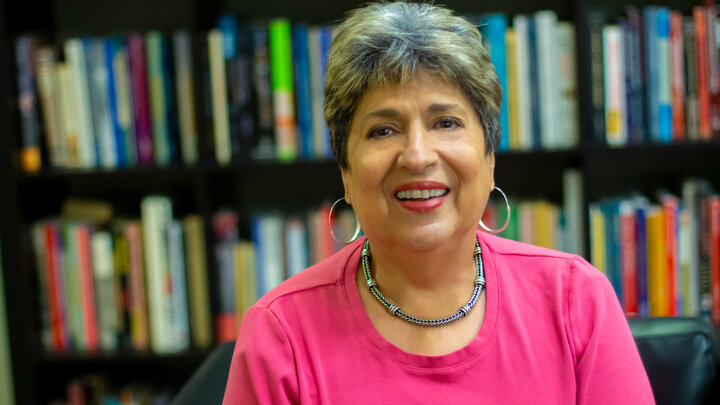 Ofelia Garcia, smiles for a photo while wearing bright pink clothing and sitting in front of book shelves. 