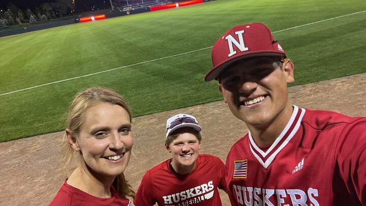 Three people, wearing red, smile for a photo with the green grass of a baseball field behind them. 