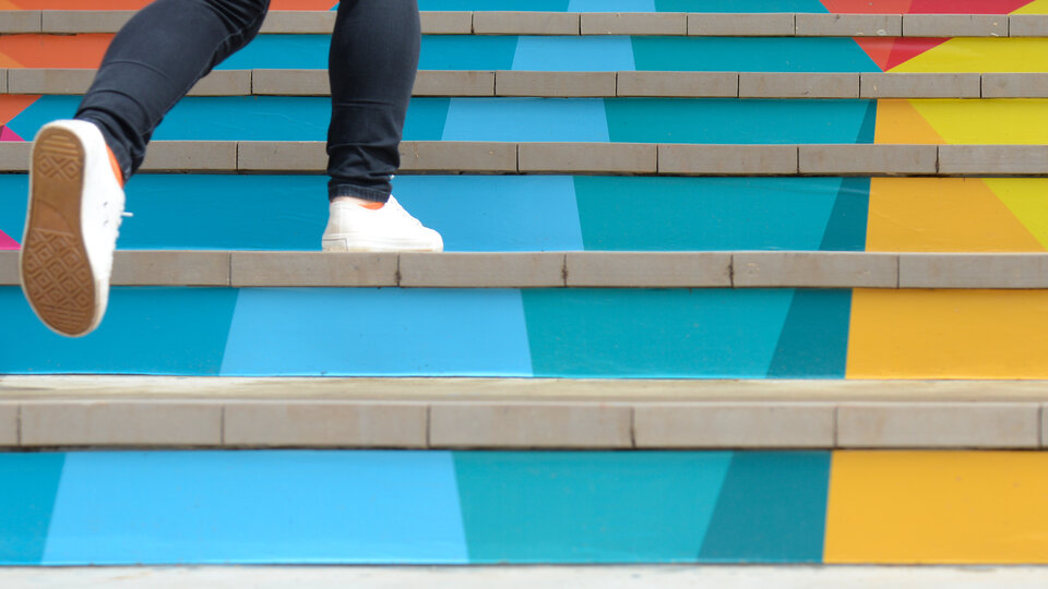 Feet in white sneakers ascending colorful stairs with geometric patterns.