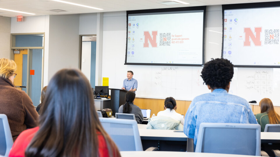 Classroom setting with a speaker at a podium and students seated, screens displaying University of Nebraska content